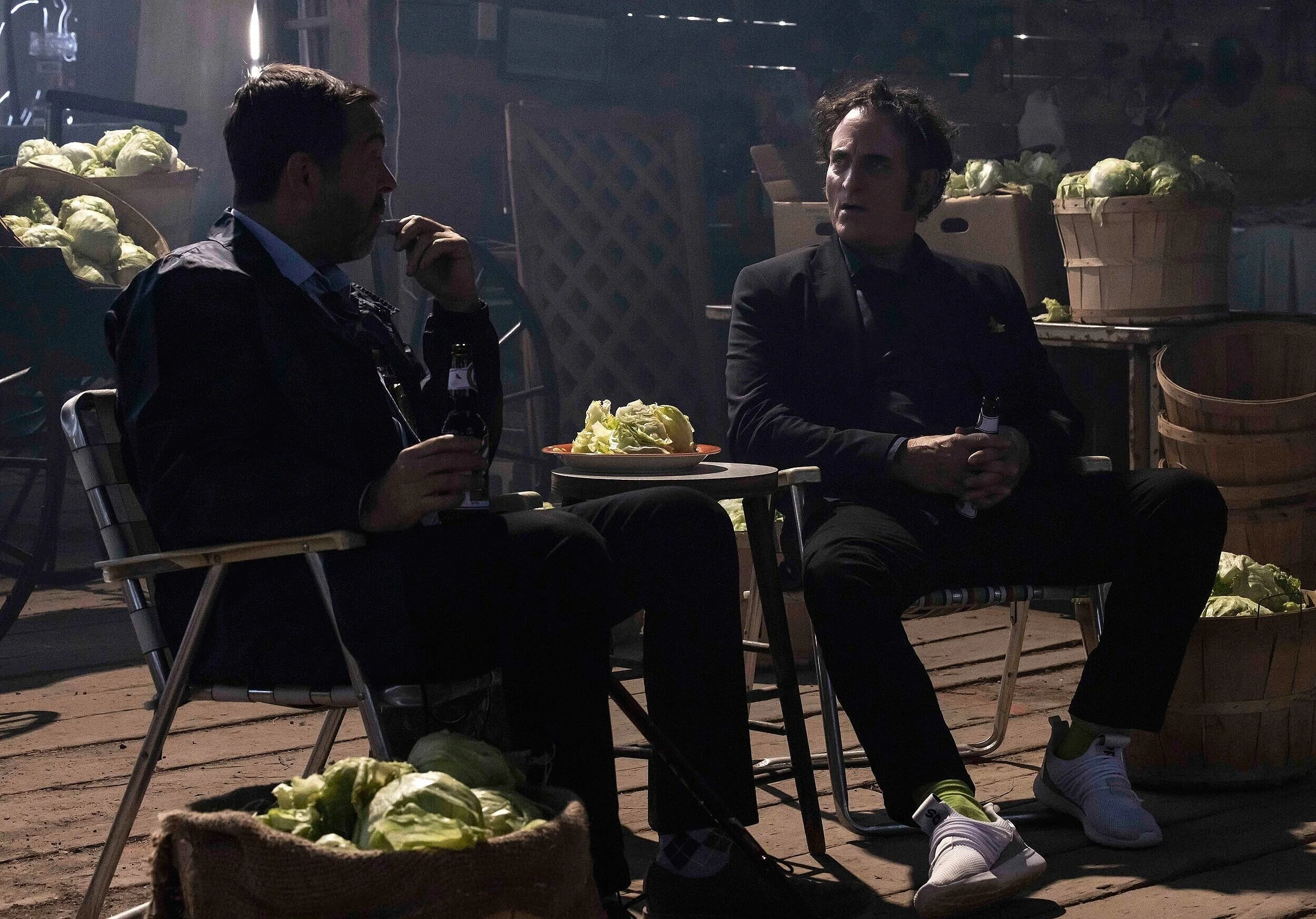 Two men sitting and talking in a dimly lit market or outdoor vegetable stand, surrounded by boxes of cabbages.