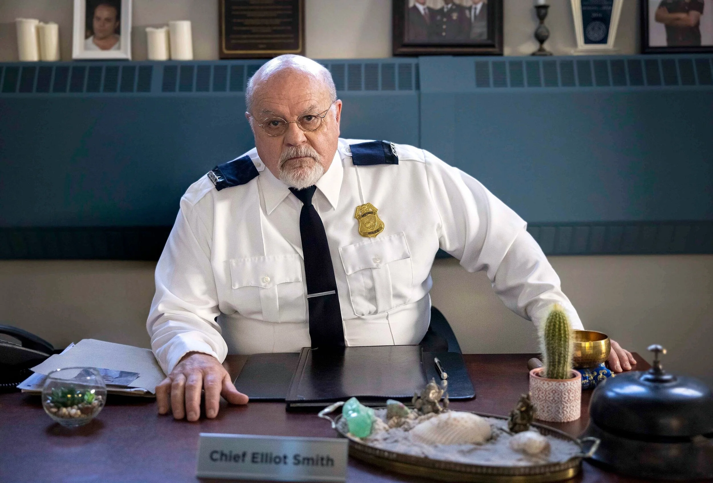 An older man wearing police uniform sitting at a desk with a nameplate that reads 'Chief Elliot Smith.' The desk has various items including a cactus, a brass bowl, a small glass bowl with decorative stones, and a black bell. The background features 