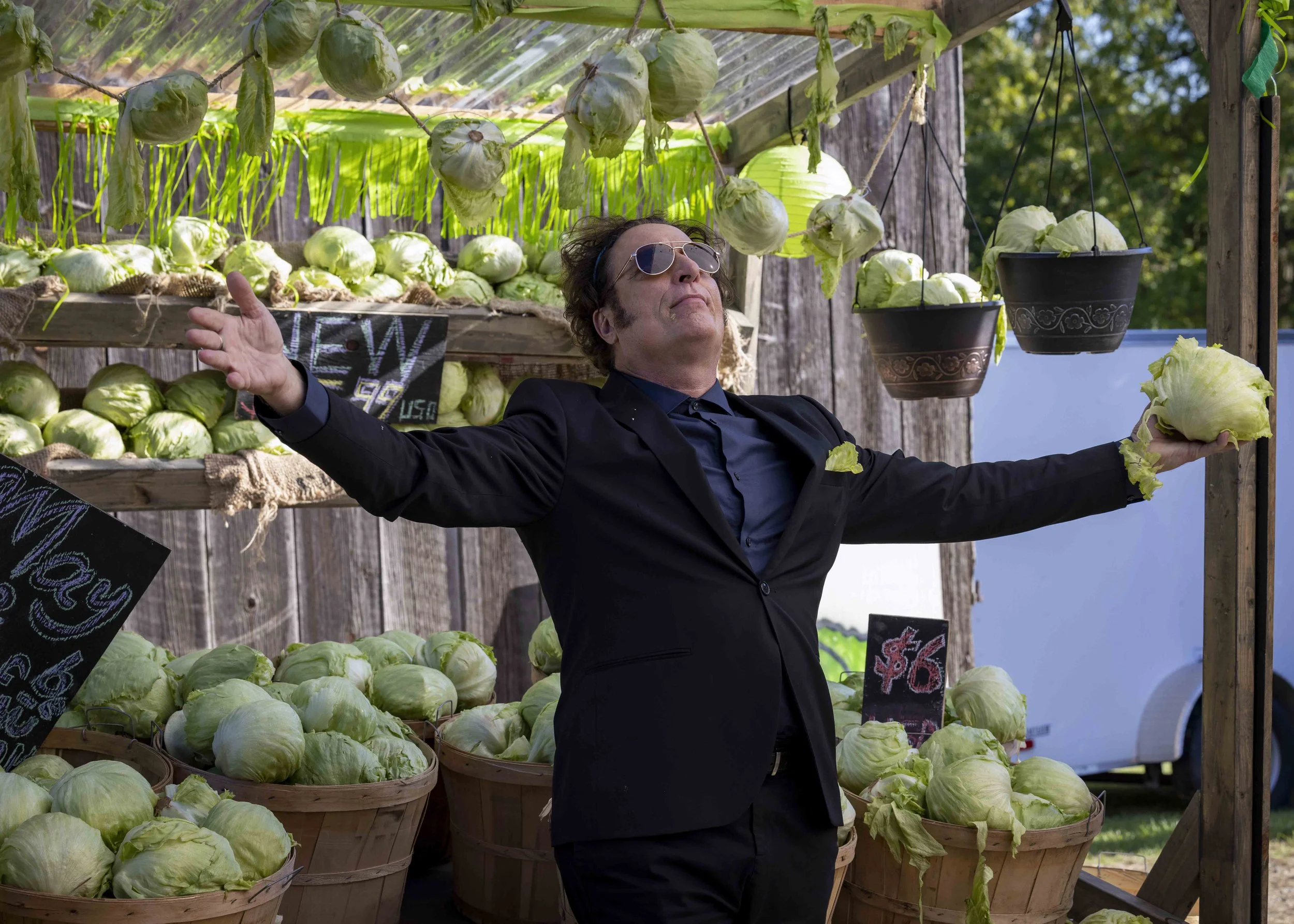 A man in a black suit and sunglasses stands with his arms outstretched in front of a stand of fresh green cabbages, with some cabbages hanging in baskets around him, at an outdoor market or farm stand.