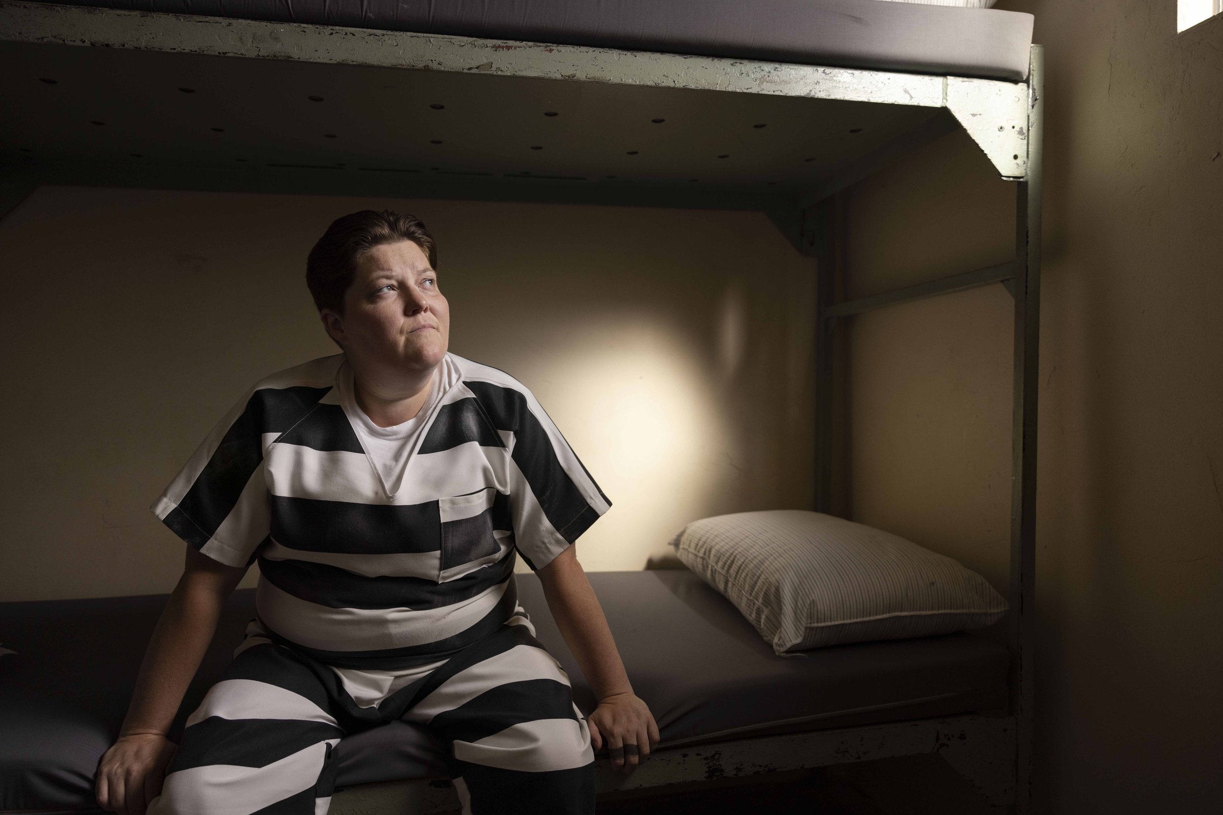 A woman in a black and white striped prison uniform sitting on a bunk bed in a dimly lit prison cell, looking thoughtfully to the side.
