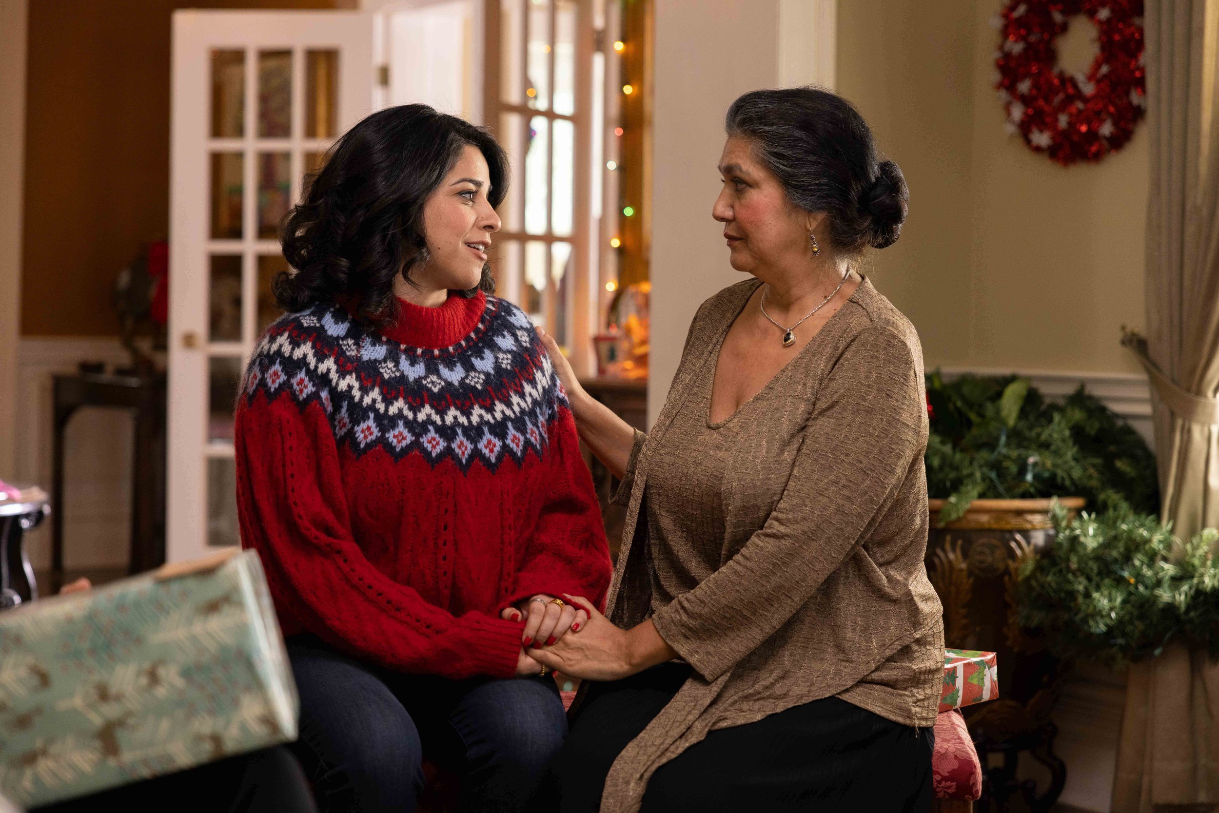 A young woman in a red Christmas sweater sitting on a chair, holding hands with an older woman who is leaning in close, in a warmly decorated room with Christmas wreath and gifts.