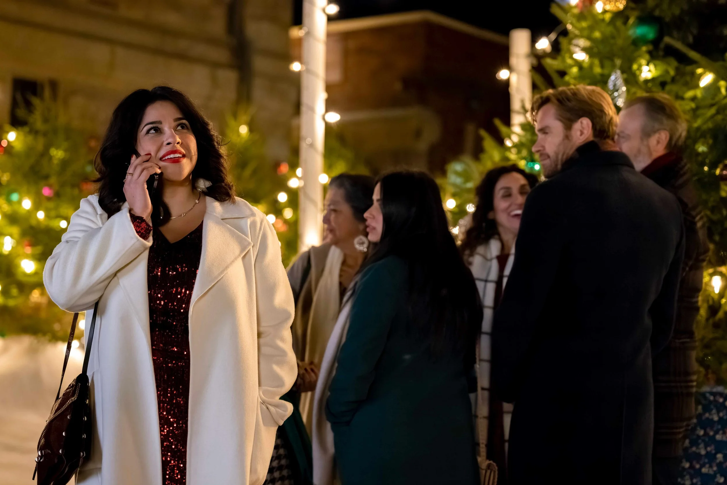 Group of people outdoors at night, decorated with Christmas lights, with a woman in a white coat talking on her phone and smiling.