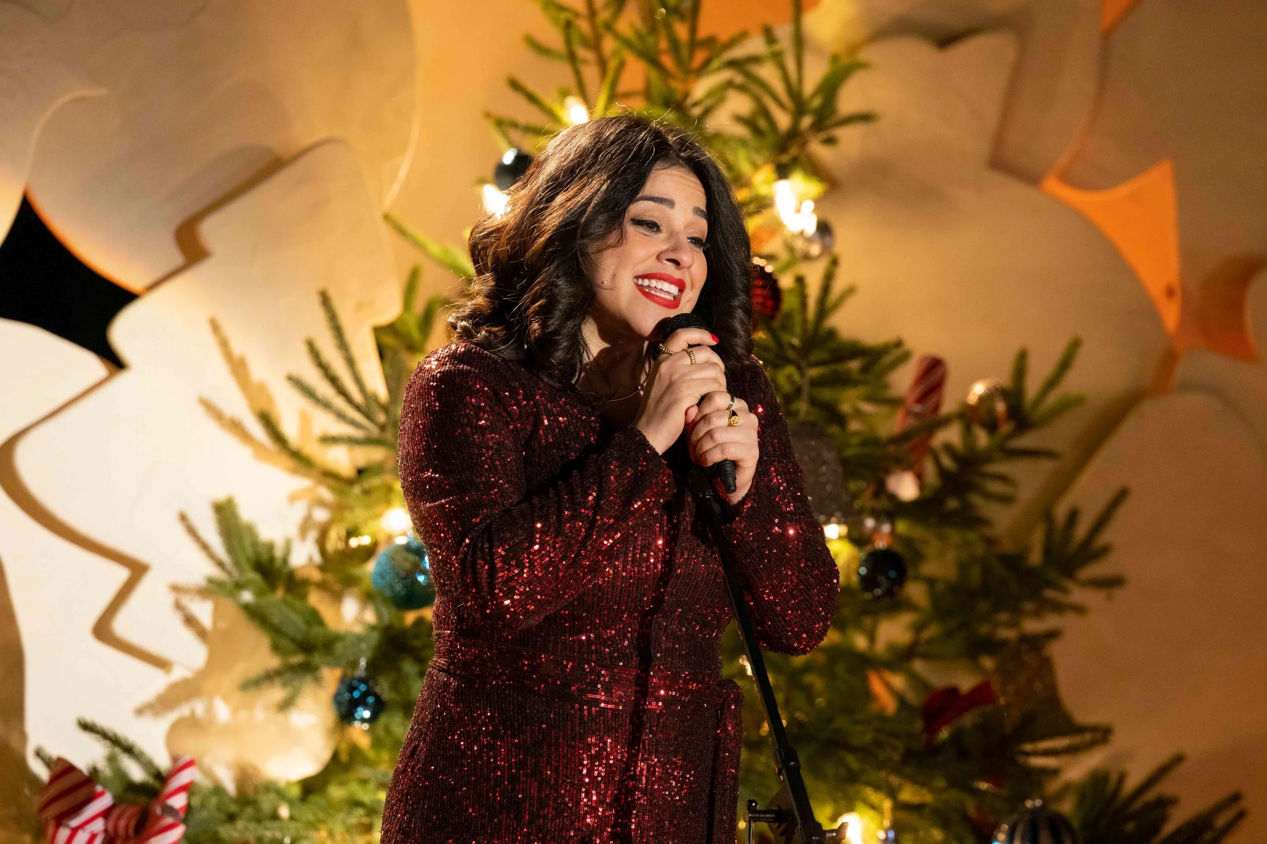 A woman with dark, wavy hair, wearing a red, sequined dress, smiling and holding a microphone in front of a decorated Christmas tree with ornaments and lights.
