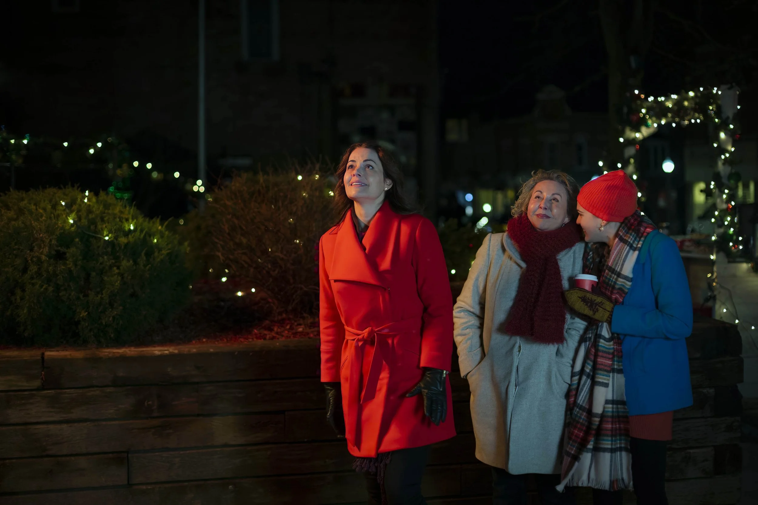 Three women standing outdoors at night during the holiday season, dressed warmly with Christmas lights and decorations in the background.