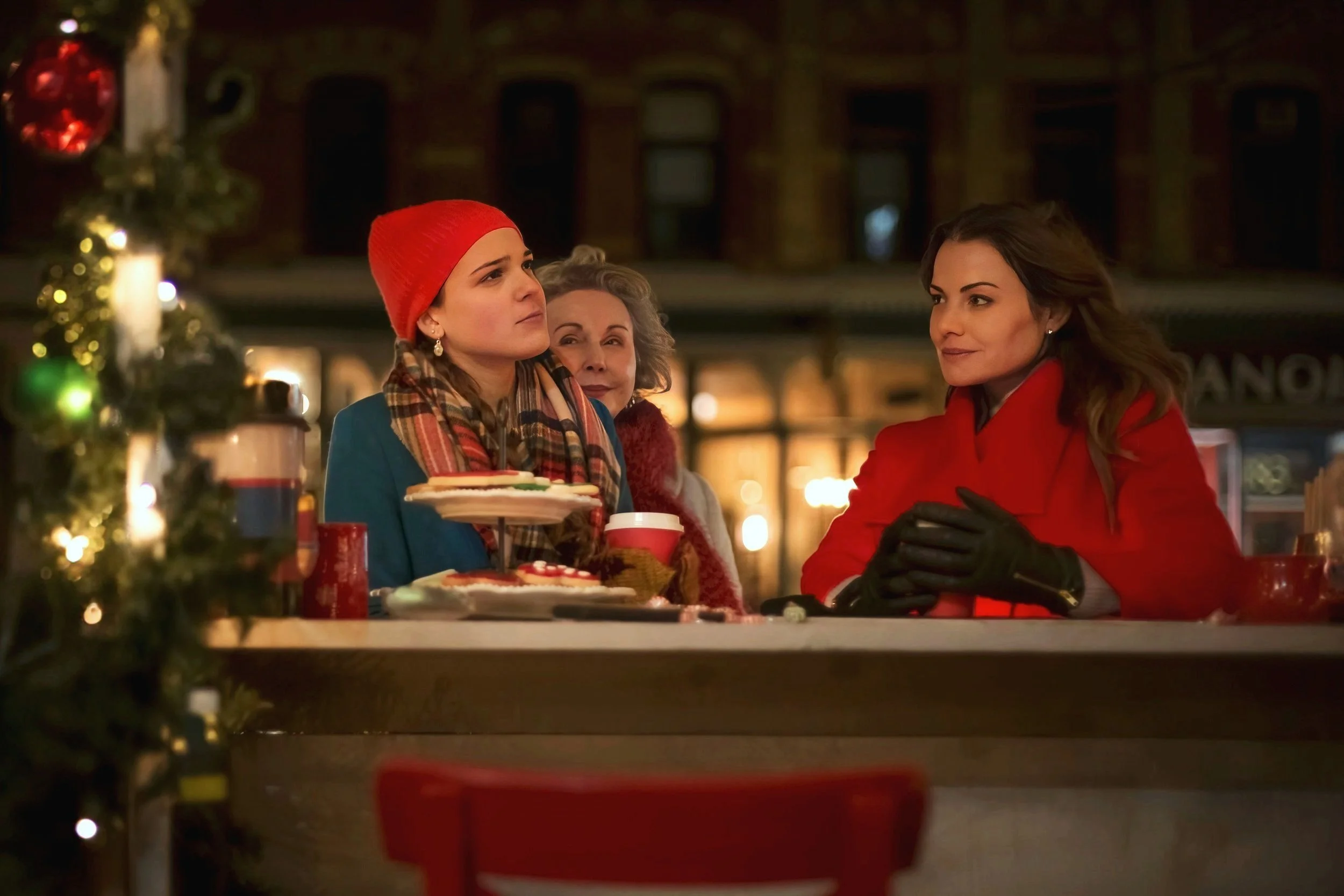 Three women sitting at a table decorated for Christmas, talking. The woman on the left is wearing a red beanie and a blue coat, the woman on the right is in a red coat with black gloves, and the woman in the middle has gray hair and is wearing a red 