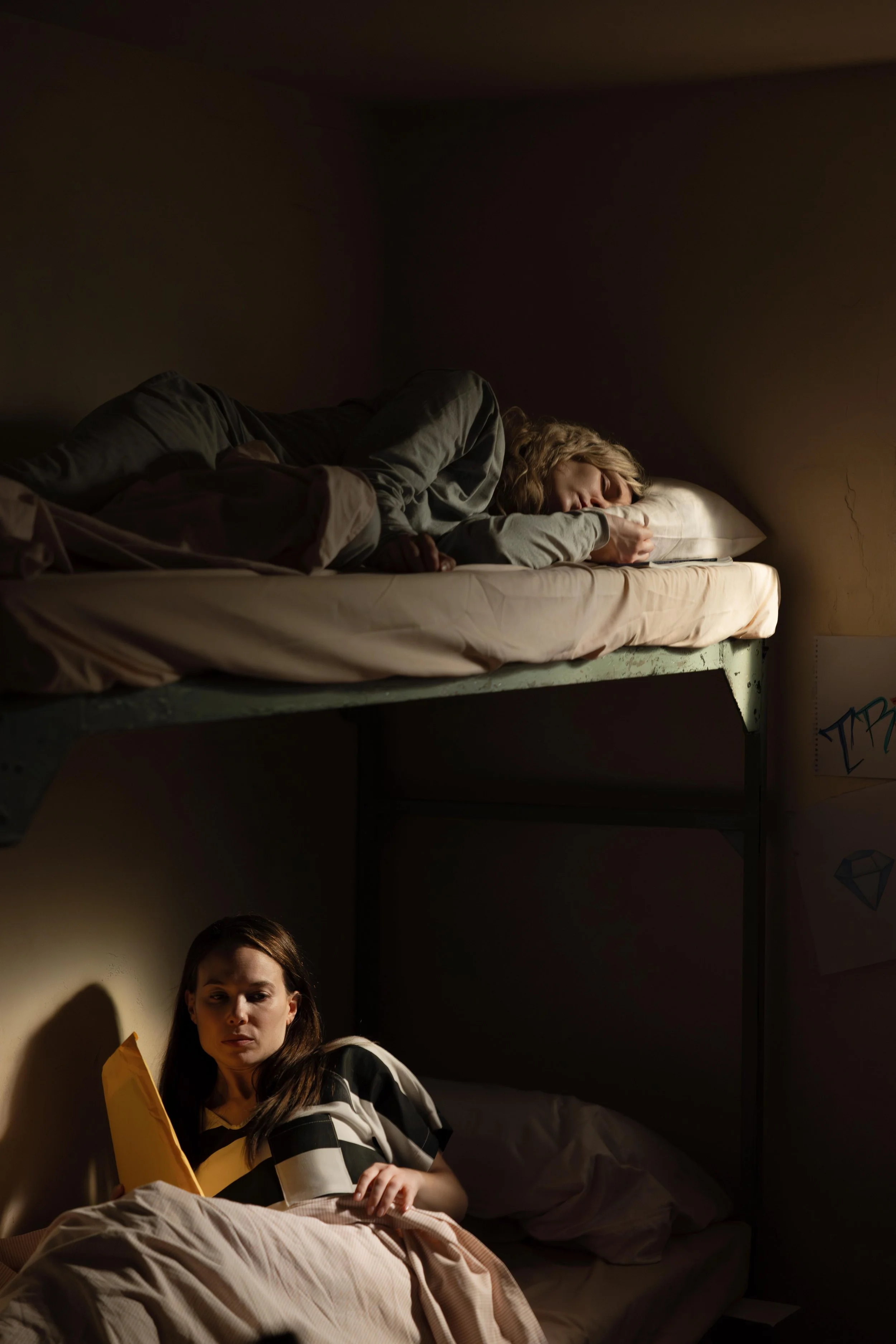 A woman lying on the bottom bunk of a bunk bed, reading a yellow envelope, with a person sleeping on the top bunk, face turned to the side, in a dark room.