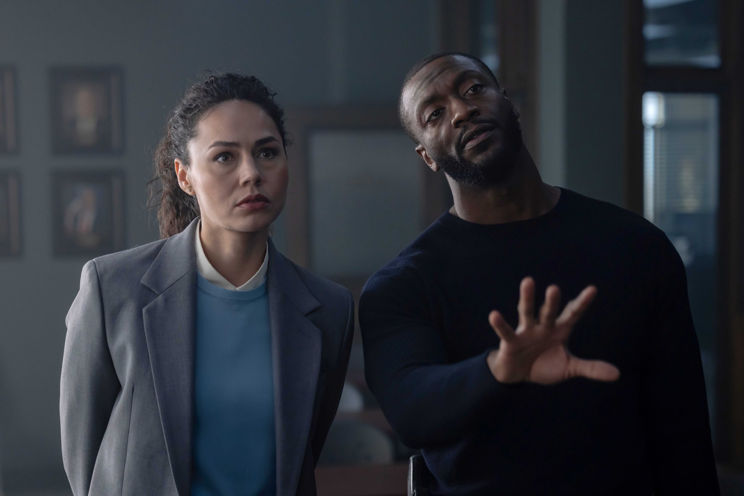 A woman and a man look at something off-camera. The woman has dark hair pulled back and wears a gray blazer over a light blue sweater. The man has a beard, short hair, and wears a black shirt, with his hand raised as if explaining or gesturing.