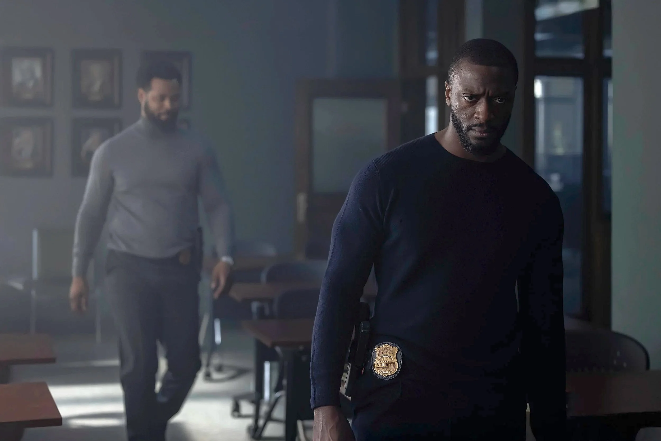 Two men, one with a police badge, in a dimly lit office or classroom, with chairs and framed pictures on the wall.