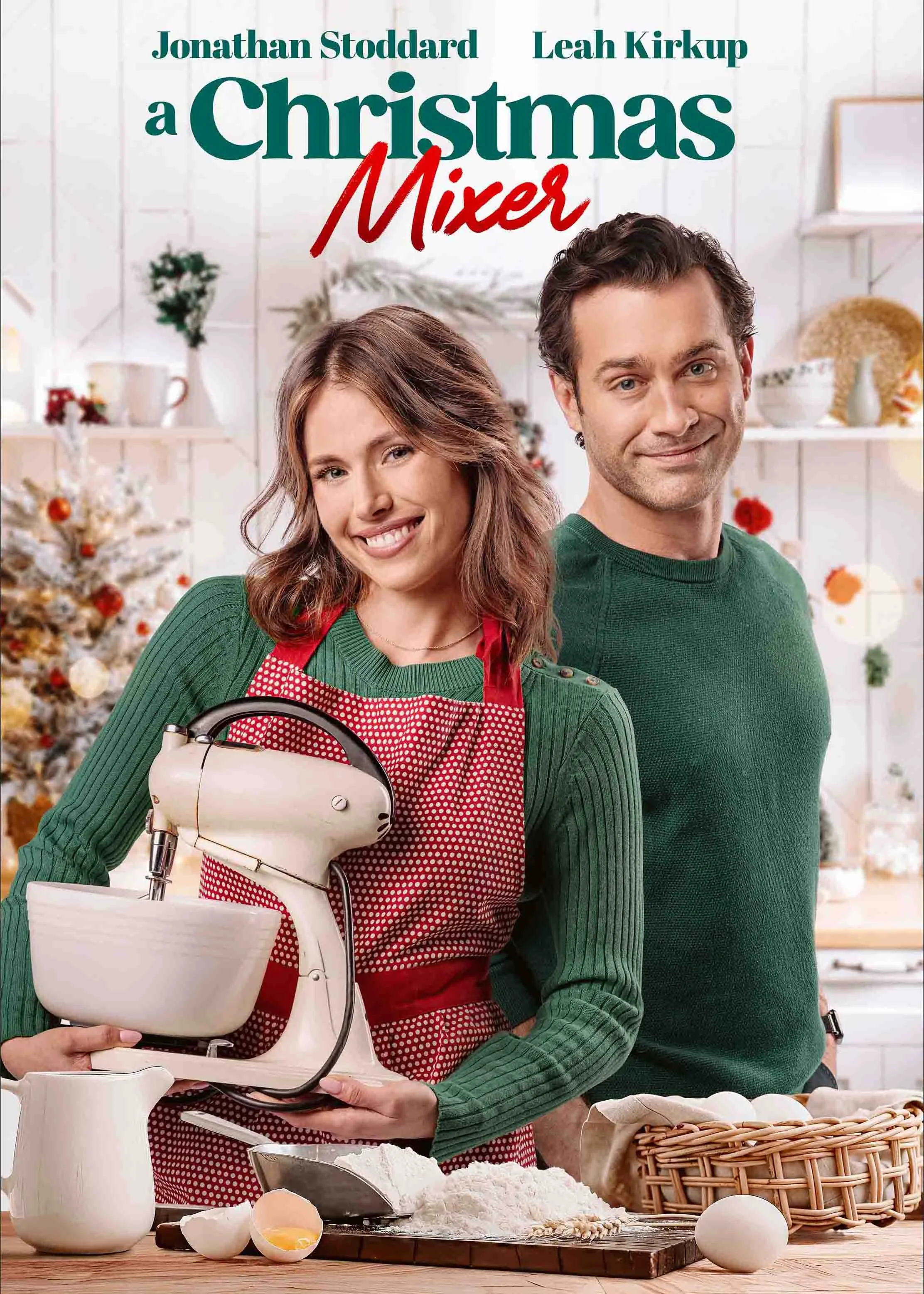 Two people, a woman in a red apron and a man in a green sweater, standing in a kitchen decorated for Christmas. The woman is holding a hand mixer and smiling. There are baking ingredients and eggs on the counter.