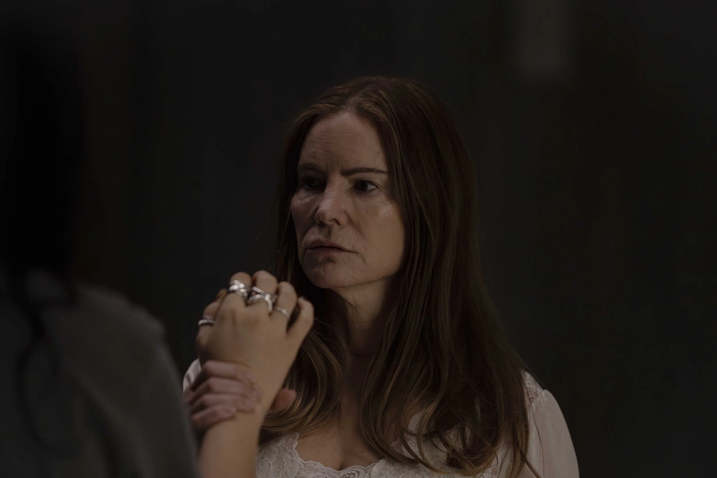 A woman with long brown hair and a serious expression, wearing a white lace top, holding hands with someone else.