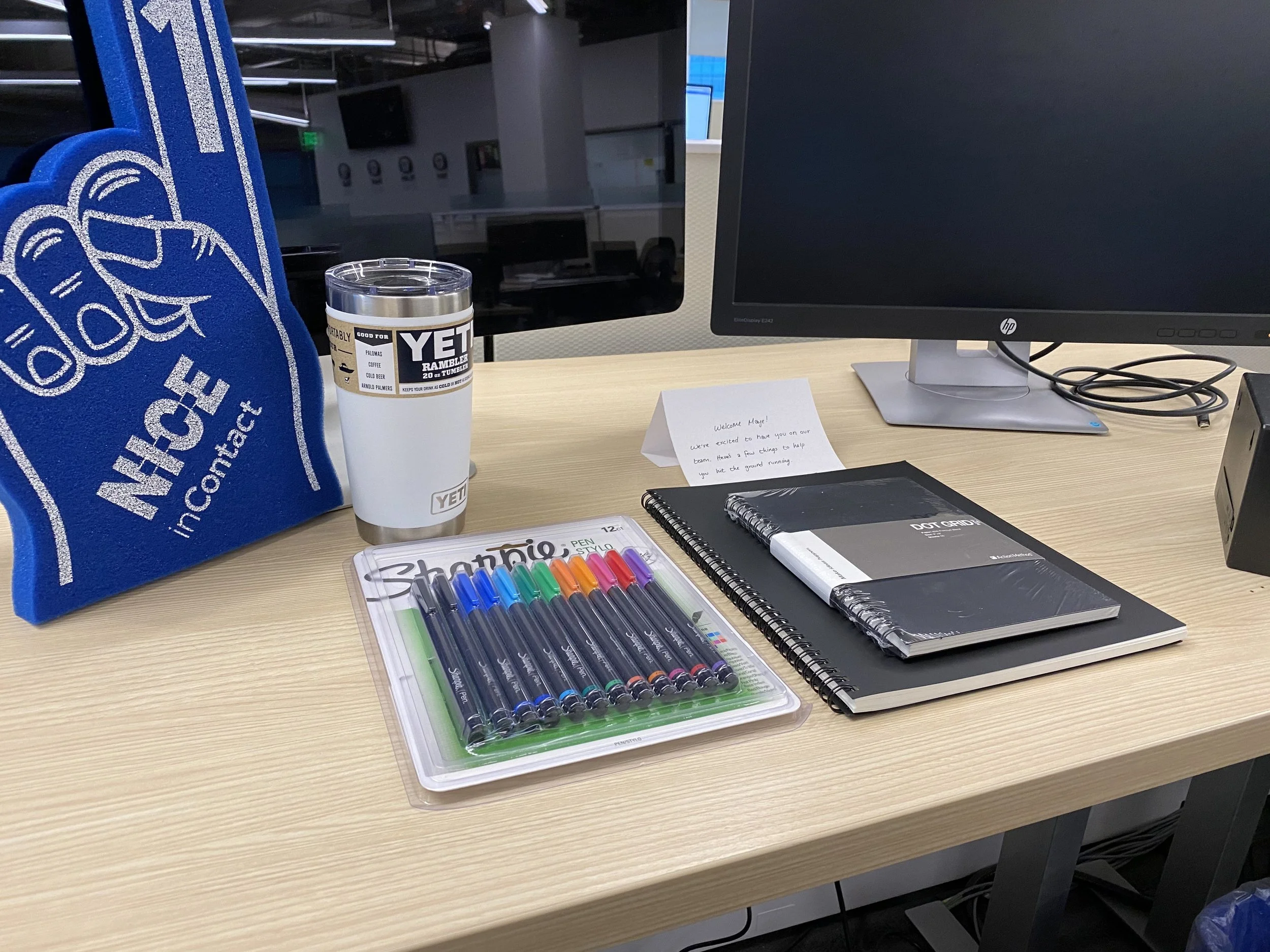 A desk setup with a blue tote bag, a YETI tumbler, three notebooks, a pack of colorful Sharpie pens, and a note saying "Welcome Maje! We're excited to have you on our team. Here are a few things to help you hit the ground running." There is a computer monitor and a keyboard in the background.
