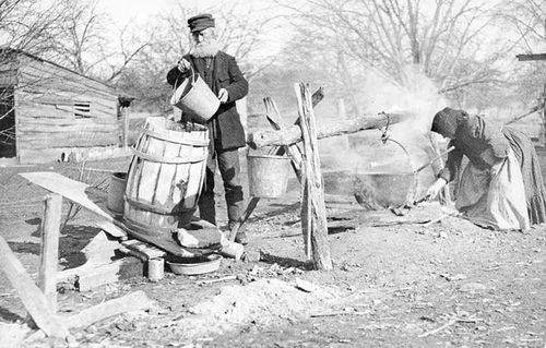 Recollections of domestic life. Soap making, 1884. The farmer is pouring water into the ash barrel to make lye; his wife is boiling a kettle of fat. Lye and fat produce soft soap.