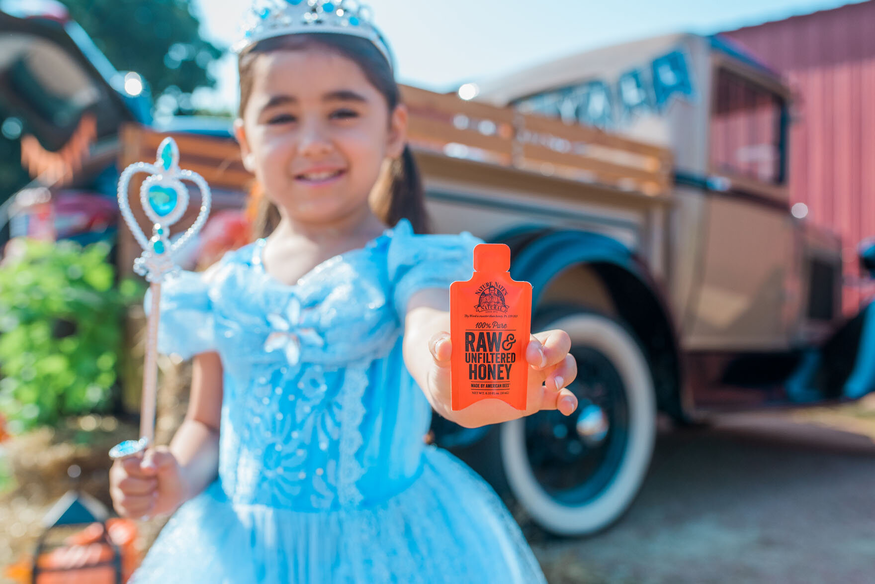Little girl in a princess costume holding a honey packet standing in front of a restored vintage truck at a fall festival