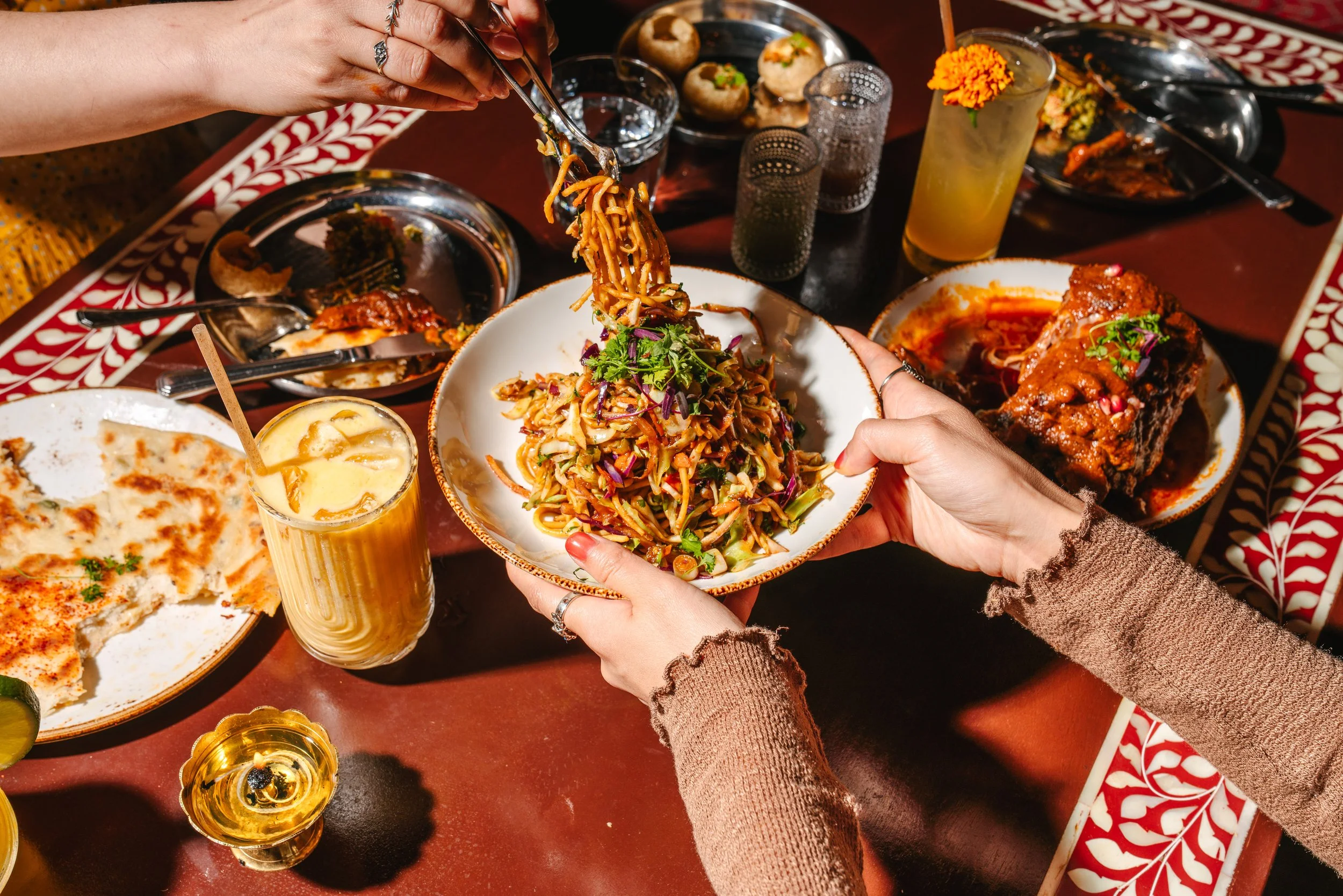 Hard light lifestyle photography in Midtown Atlanta for Ghee Indian restaurant showing people sharing a meal with various Indian dishes such as noodles, naan, and curry, along with drinks on a decorated table.