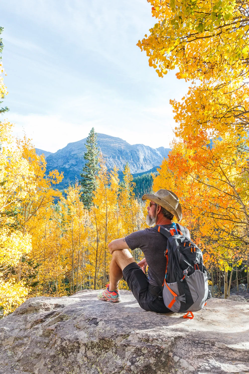 An older man with a gray beard wearing a cowboy hat, glasses, a gray t-shirt, and shorts, sitting on a large rock in an autumn forest with yellow and orange leaves. He has a backpack and is gazing at distant mountains.