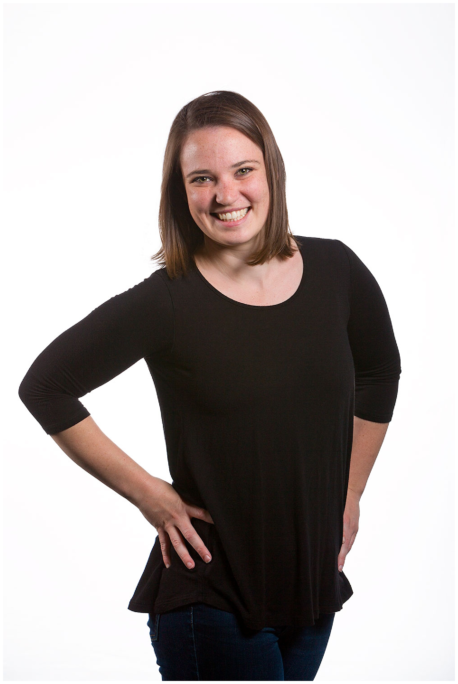 A young woman with shoulder-length brown hair, smiling, wearing a black top with three-quarter sleeves and dark jeans, standing with one hand on her hip against a plain white background.