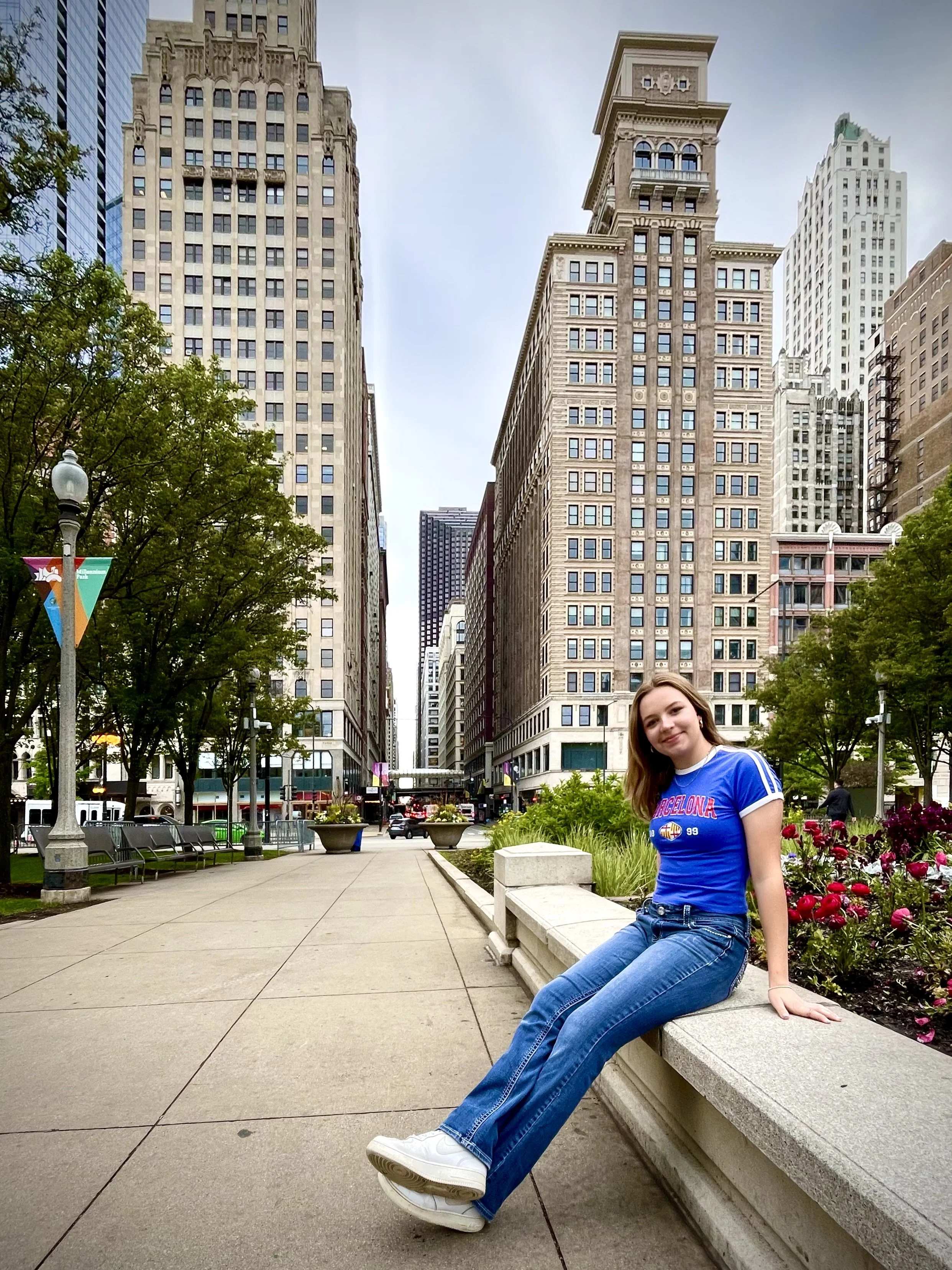 A young woman in blue jeans and a blue t-shirt sitting on a stone bench in a city park with tall buildings and trees in the background.