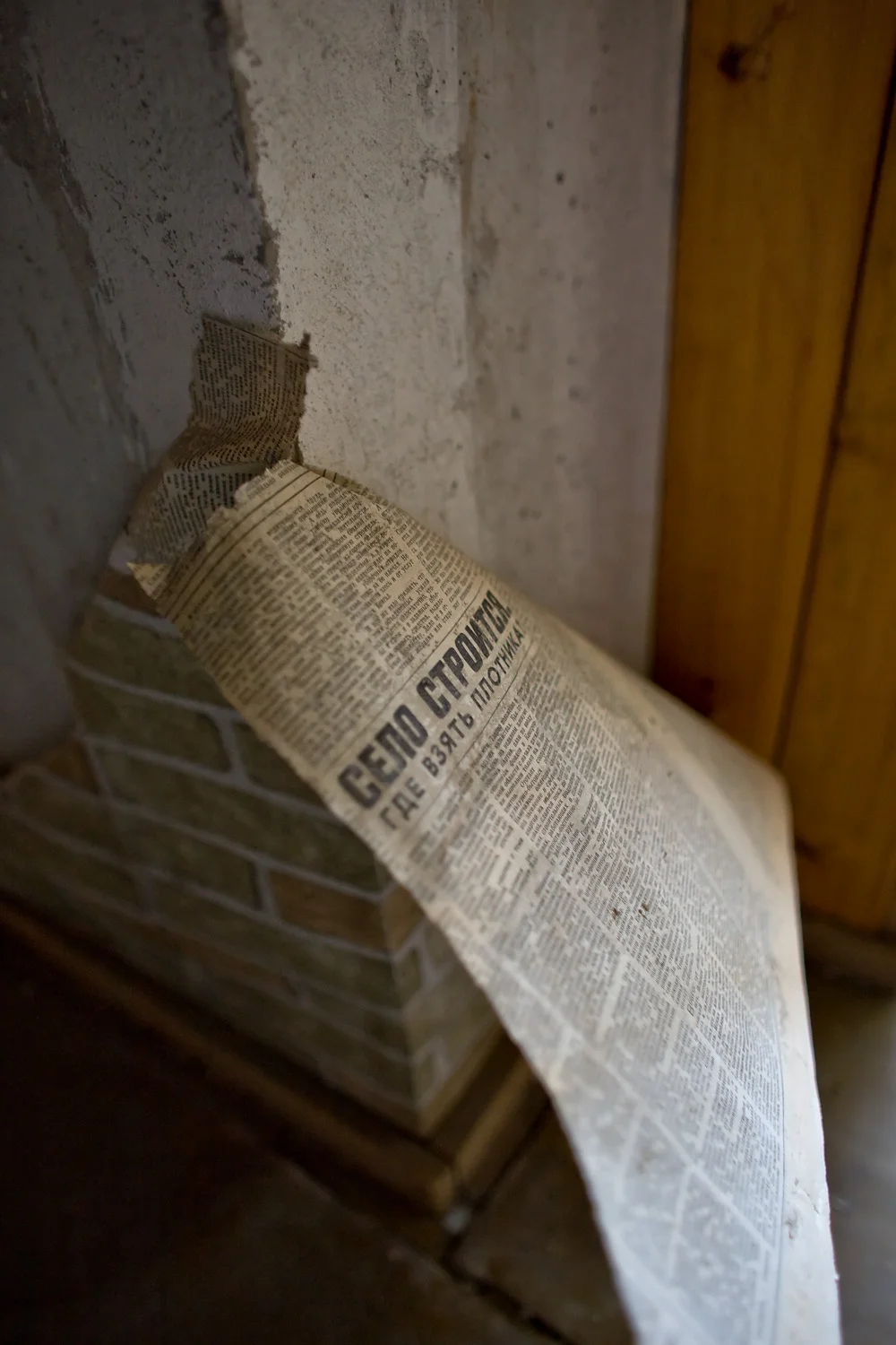 Peeling fake brick wallpaper, showing the newspaper backing. The title of the article reads "Village construction - Where to find carpenters?"