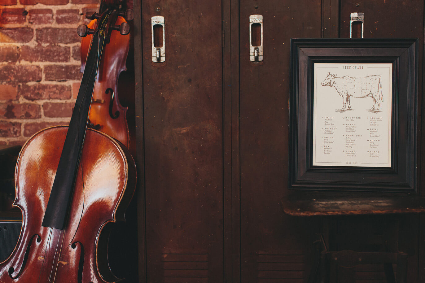 Hand drawn illustrated fine art print diagram of types of cuts of beef is framed in a dark wood frame and sits in front of lockers and next to an old violin.