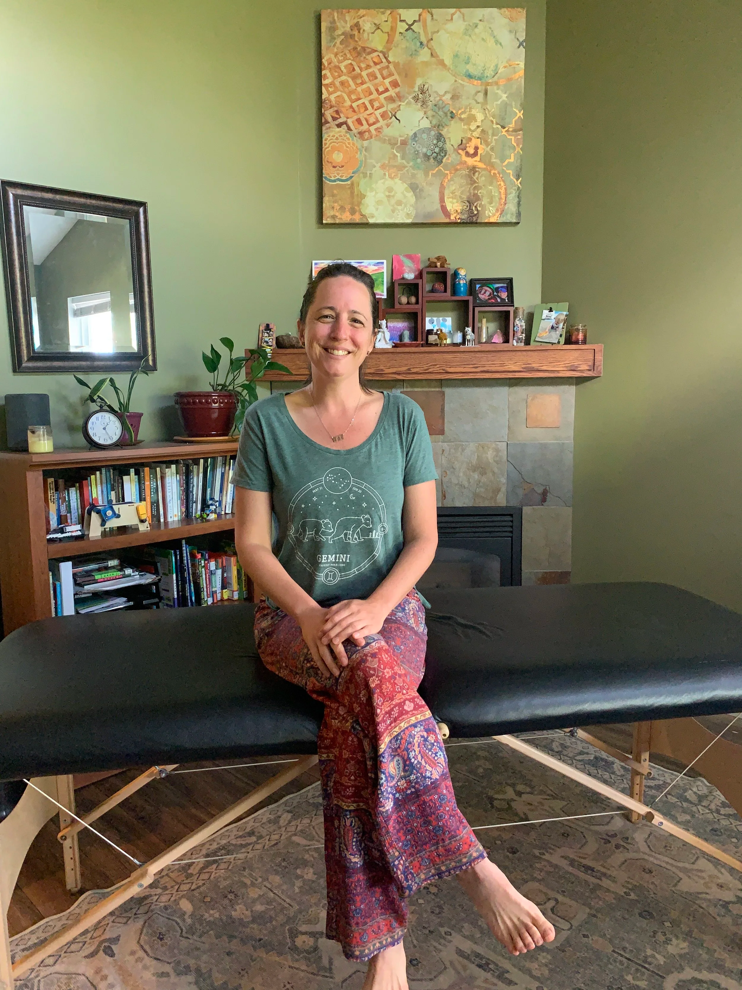 Beverly Hosford sitting on a massage table in a living room, smiling at the camera, with a fireplace, a bookshelf, plants, and artwork in the background.