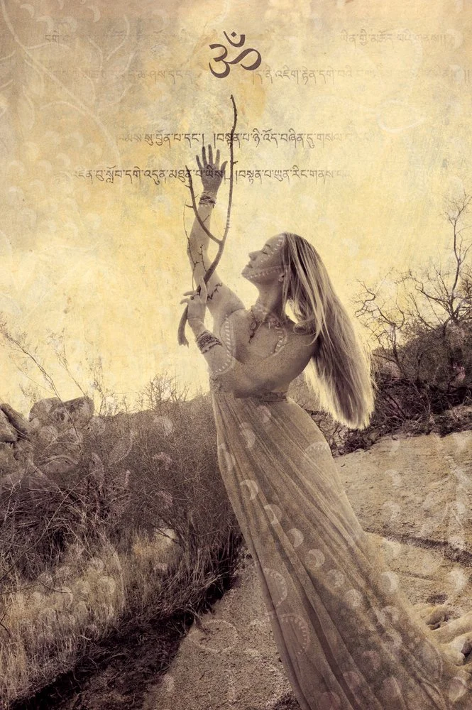 A woman with long blonde hair wearing traditional clothing, holding a bow and arrow, standing outdoors in a desert landscape with dry bushes and rocks.