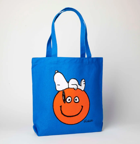 A bright blue canvas tote bag with a screen-printed illustration of Snoopy lying happily on a large orange smiley face.