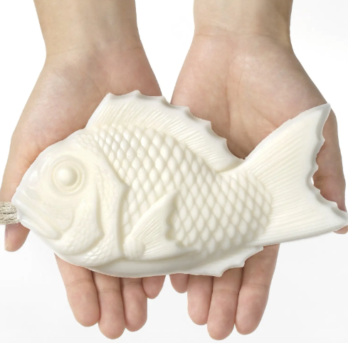 A pair of hands holding the fish-shaped Tamanohada Welcome Gift Soap by Ameico, showcasing its detailed scale texture.