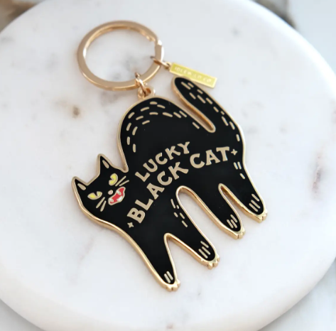 The Lucky Black Cat enamel keychain resting on a white marble surface, highlighting its polished brass finish and vibrant enamel details.