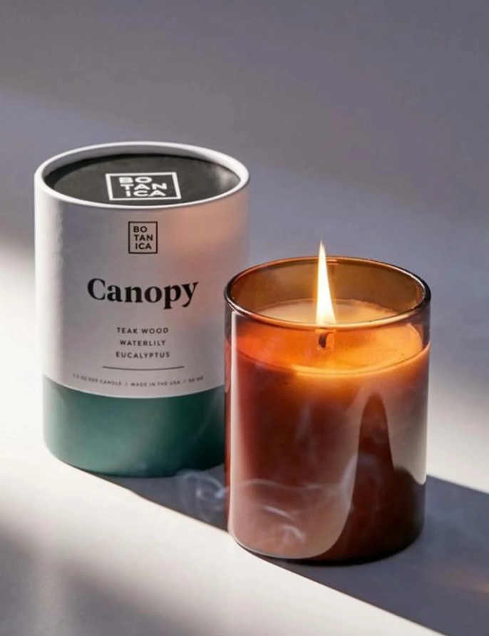 Canopy Candle by BOTANICA in a glass container with the packaging box, lit to reveal a warm glow.