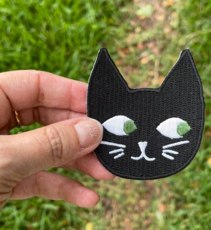 A 3-inch embroidered patch of a smiling black cat with bright green eyes.