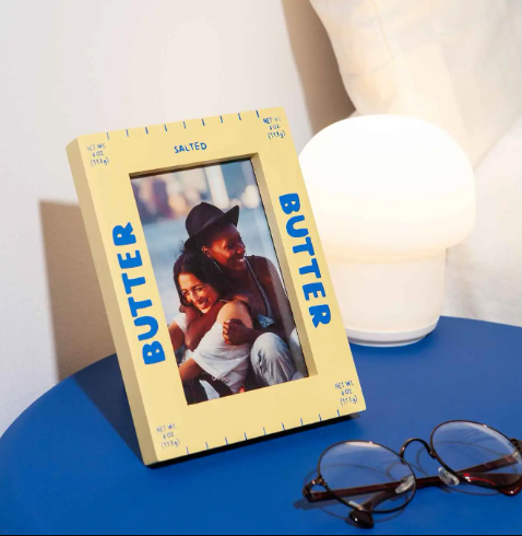 The yellow butter photo frame displayed on a blue table with a photo inside, showcasing its unique and playful appeal as home decor.