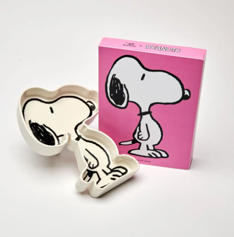 The Magpie Peanuts Snoopy Shaped Trinket Dish, made of white ceramic, displayed next to its vibrant pink gift box featuring a bold illustration of Snoopy.