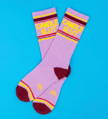 Display your witty style with the lavender Candle Slut gym crew socks by Gumball Poodle, featuring bold yellow lettering and striped accents against a bright blue background.