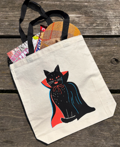 Style your everyday adventures with the Vampire Cat Neon Screenprinted Tote by Zander Mack, conveniently packed with daily essentials on a rustic wooden surface.
