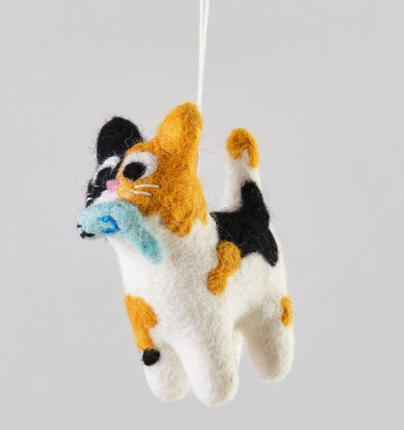 A white, black, and orange calico cat felted wool ornament carrying a light blue fish in its mouth.