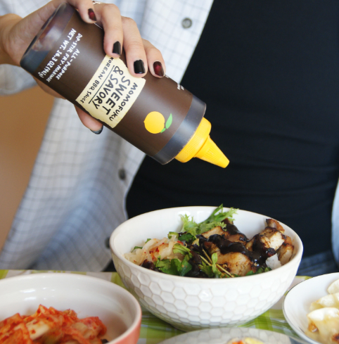 A person drizzling Momofuku Sweet & Savory Korean BBQ Sauce over a fresh rice bowl with grilled chicken and crisp greens.