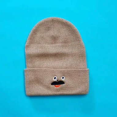 Kristina Micotti's humorous Dad Beanie, a tan knit hat featuring an embroidered mustache face design perfect for gifting.