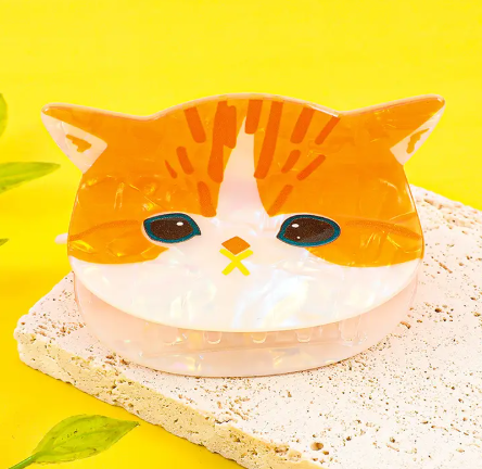 A whimsical hair claw clip shaped like an orange and white tabby cat face with a glossy acrylic finish.