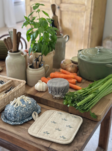 A kitchen scene featuring the embroidered linen pot holder on a wooden cutting board surrounded by various cooking ingredients and stoneware.