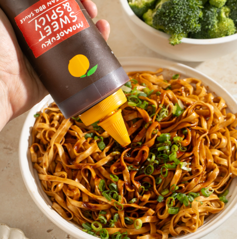 A hand squeezing Momofuku Sweet & Spicy Korean BBQ Sauce onto a delicious bowl of stir-fried noodles topped with scallions.
