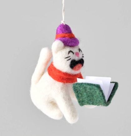 A white felted wool cat ornament wearing a red scarf and purple hat, singing from a small green songbook.