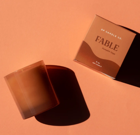 The Fable 9 oz. Hue Candle by GP Candle Co. sits alongside its matching brown gift box on a warm terracotta surface.