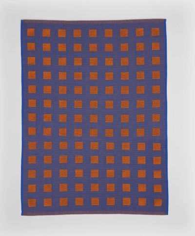 A 100% woven cotton tea towel with a graphic square pattern in contrasting burgundy and blue colors, laid out flat.