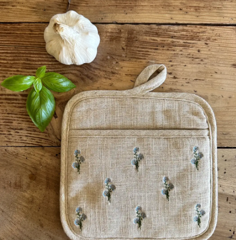 A close-up shot of a natural linen pot holder with blue, green, and yellow floral embroidery, resting on a rustic wooden table next to fresh basil and garlic.
