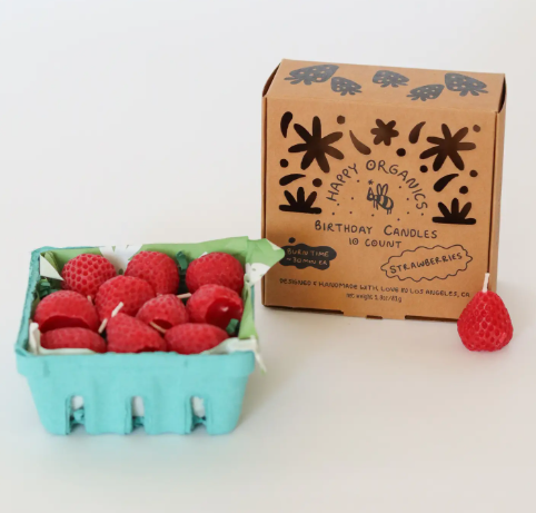 Present a lovely retail setup with a teal paper pulp basket filled with textured strawberry-shaped candles next to a brown kraft box featuring the Happy Organics.