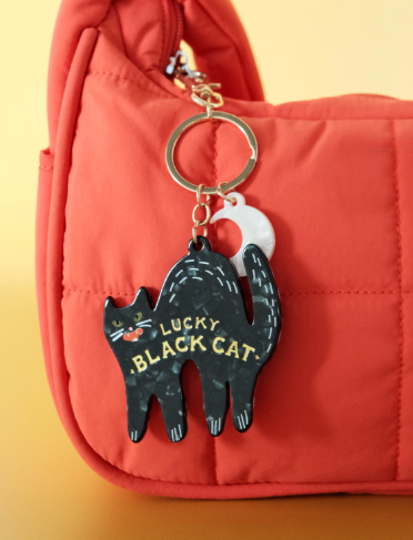 A black cat-shaped Lucky Bag Charm with gold lettering, featuring a moon charm, attached to a vibrant orange bag.