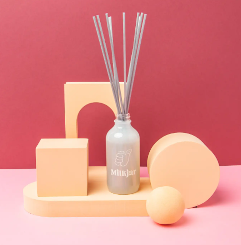 Milkjar's Dandy reed diffuser in a frosted glass bottle with its reeds fanned out, artfully arranged with geometric shapes against a vibrant red and pink background.