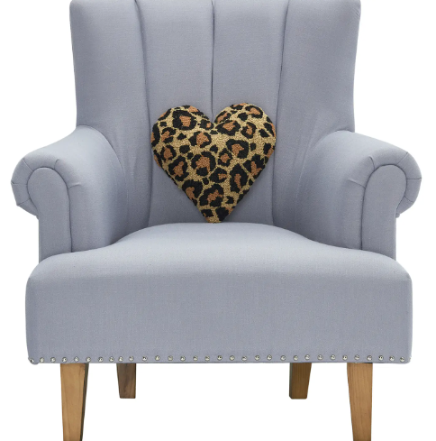 The Peking Handicraft Leopard Heart Hook Pillow placed centrally on a stylish, light-colored upholstered armchair, highlighting its unique heart-shaped silhouette and animal print design.