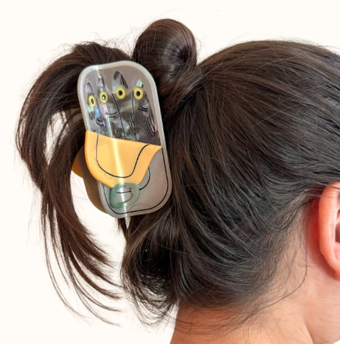 A person's dark hair styled into a messy bun, secured with the large tinned fish hair claw clip, showcasing its size and playful detail.