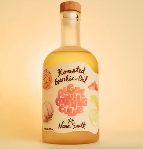 Highlight the bottle of Nara's Roasted Garlic Oil by Algae Cooking Club on a kitchen prep surface alongside whole garlic bulbs and a freshly baked flatbread.